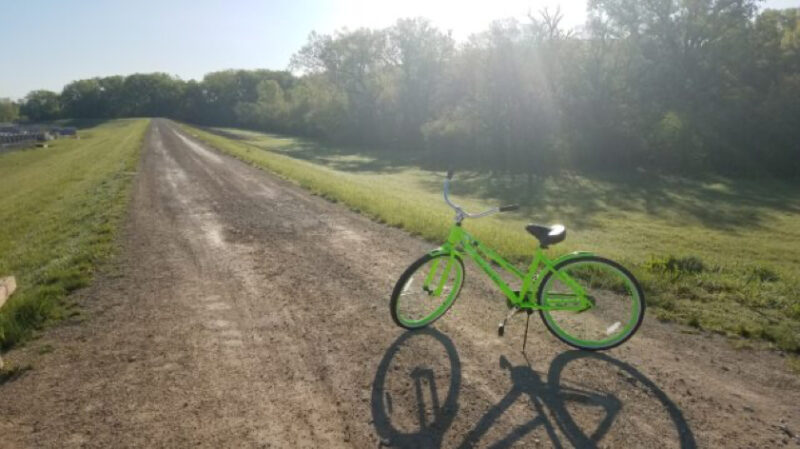 Photo of a bikeshare bike on its kickstand on a dirt trail surrounded by grass and trees